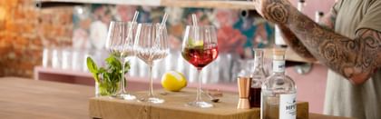 NACHTMANN Gin and Tonic glasses with glass straws on a bar counter. One is filled with a red Gin Cocktail with mint and a lemon zest. Behind the glasses is a lemon and a glass with mint sprigs. On the right are the hands of the bartender and bar equipment.<br/>
