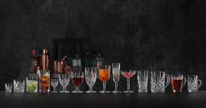 The NACHTMANN Noblesse collection lined up on a black bar counter, in the background are bottles and bar equipment. The Noblesse tumbler, longdrink glass, cocktail glass and coupette are filled with different cocktails. The wine goblet is filled with red wine, the hot beverage mug is filled with tea. The other glasses are empty.