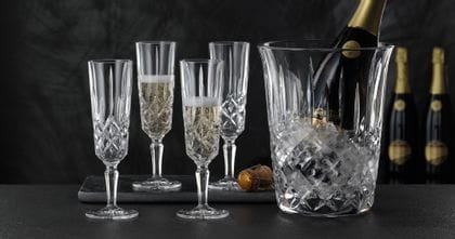 Four NACHTMANN Noblesse Champagne glasses two of which are filled with Champagne. Next to them is the NACHTMANN Noblesse Champagne cooler with ice cubes and an open bottle of Champagne inside.<br/>