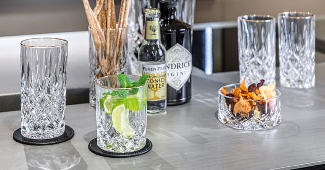 The NACHTMANN Noblesse Gold longdrink glass next to the tumbler filled with a gin and tonic cocktail. In the background one more longdrink glass filled with crissini and a bottle of tonic water.<br/>