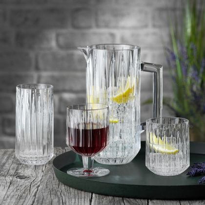 The NACHTMANN Jules crystal glass series on a round serving tray. The pitcher and the tumbler are filled with water, ice cubes and lemon slices, to their right is lavender. The goblet is filled with red wine. The longdrink glass in the background is empty.<br/>