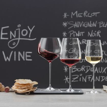 SPIEGELAU Willsberger Anniversary glasses on a marble sideboard. On the left an empty Bordeaux glass and white wine glass, in the middle a pile of corks and potato chips, on the right the filled Burgundy glass, filled red wine glass and filled white wine glass. In the background a blackboard with a menu and the words drink wine.<br/>