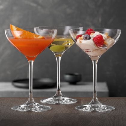 SPIEGELAU Dessert and Champagne saucers on a wooden table, each of them filled with a different cocktail drink and one with a dessert with berries on it.<br/>