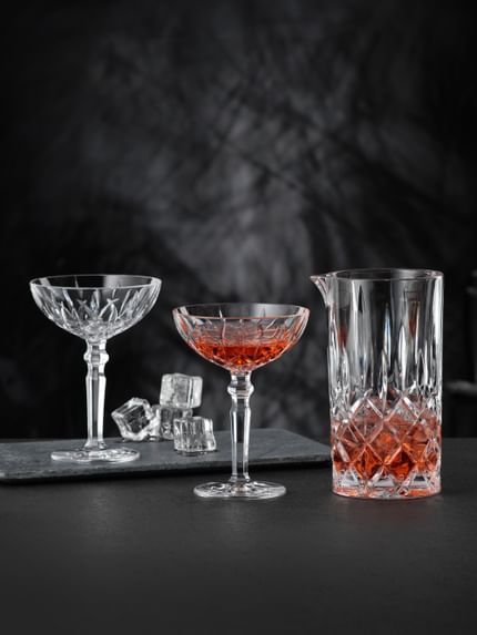 The NACHTMANN Noblesse pitcher filled with a red drink and ice cubes and two NACHTMANN Noblesse cocktail glasses, one of them filled with the same red drink. In the background there is the empty cocktail glass and ice cubes.<br/>